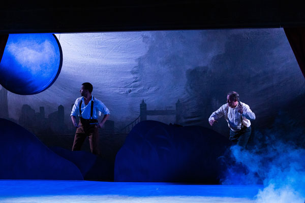 stage scene showing two men on a misty stage in shades of deep blue with a large blue moon in the background, one man holds a gun; both look offstage left