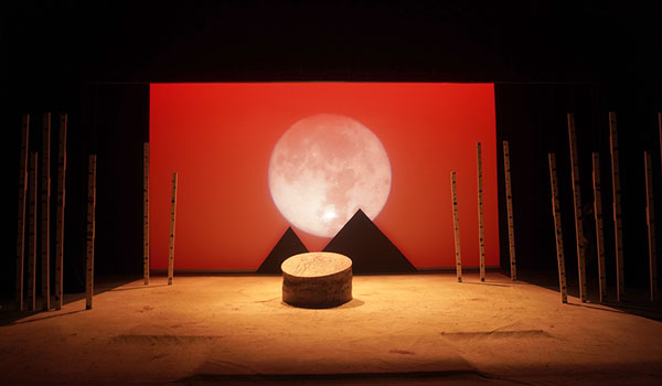 an empty stage with a red backdrop featuring a large white moon and two pyramid silhouettes, with tall sticks around the edge of the stage and a round table in the center