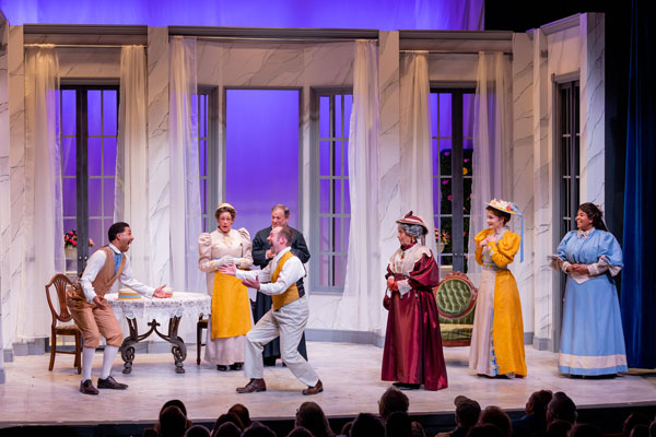 stage scene showing three men and four women in early 1900s style clothing in front of open windows; the men face each other with expressions of excitement