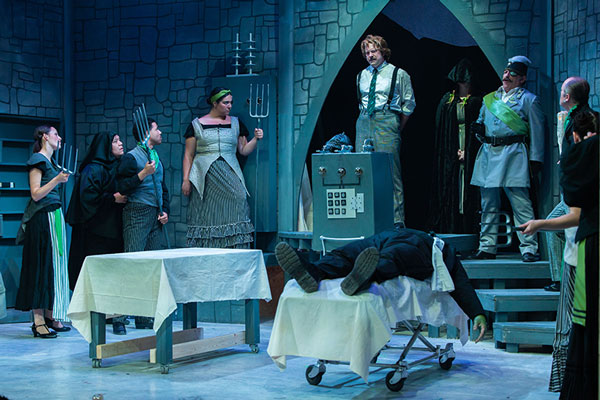 stage scene showing castle interior, with science lab equipment, a Frankenstein monster lying on a gurney, villagers holding pitchforks