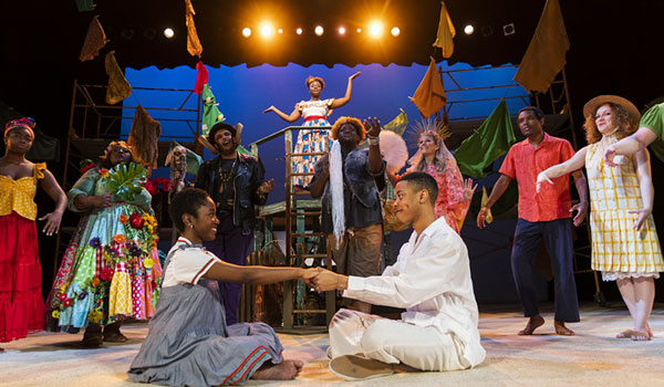 colorful cast of characters in island garb on stage, with a young black man and woman sitting front and center, holding hands and smiling
