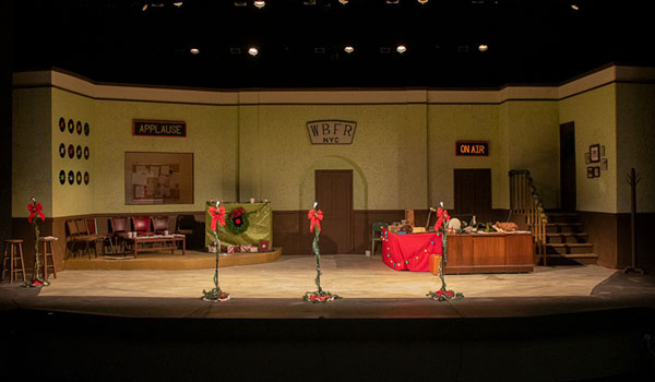 an empty stage decorated with vintage furniture, Christmas decorations, and three microphones on stands at the front