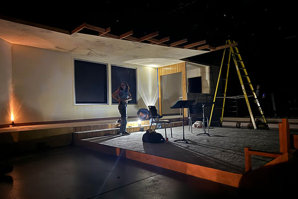 process photo showing a stage in the early stage of being built; a young woman stands near a ladder in front of open windows