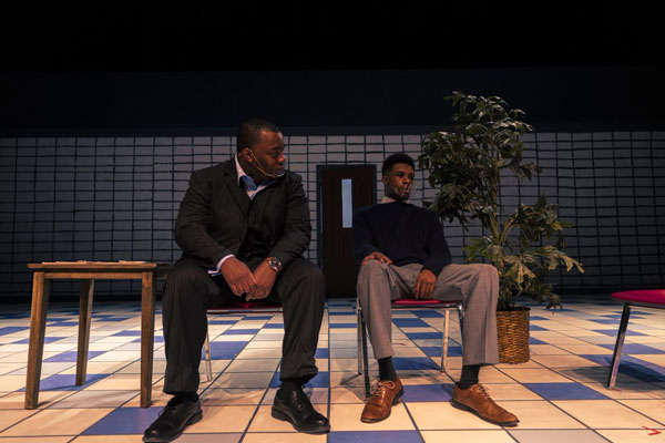 An older and younger black male actor sit in chairs onstage