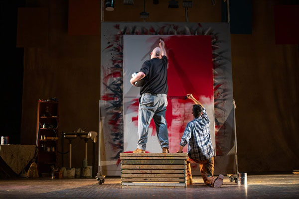 Two young male actors painting a large canvas with backs to the camera