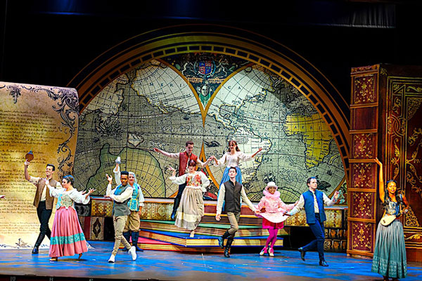 stage scene showing many young women and men in costumes that evoke fairy tales, the background is a huge vintage world map, and there are oversized books and pages comprising the scene as if the whole stage is a book brought to life